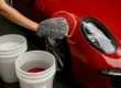 Foto detalhada de uma pessoa lavando um carro esportivo vermelho brilhante na sombra, utilizando uma luva de microfibra e a técnica dos dois baldes com um separador de partículas (Grit Guard) visível no balde de enxágue.