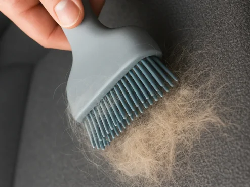 Close-up de uma escova de cerdas de silicone sendo usada para remover uma grande quantidade de pelos de cachorro (ou gato) presos no carpete cinza do banco de um carro.