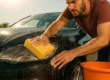 Foto ilustrativa mostrando um homem lavando um carro preto sob sol forte, usando um balde único e uma esponja amarela de cozinha, simbolizando os erros comuns que levam a microrriscos e danos na pintura automotiva.