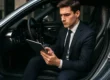 Um motorista jovem e engravatado está sentado no banco de um carro esportivo com a porta aberta, utilizando um tablet para analisar atentamente um contrato de seguro automotivo (apólice)