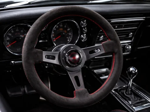 Interior de um carro com volante esportivo de três raios instalado, destacando o acabamento em camurça e costuras vermelhas.