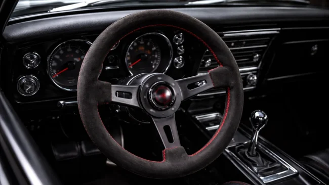 Interior de um carro com volante esportivo de três raios instalado, destacando o acabamento em camurça e costuras vermelhas.