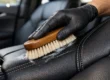 Pessoa utilizando escova de cerdas macias para limpar detalhadamente um banco de couro preto de um carro de luxo.