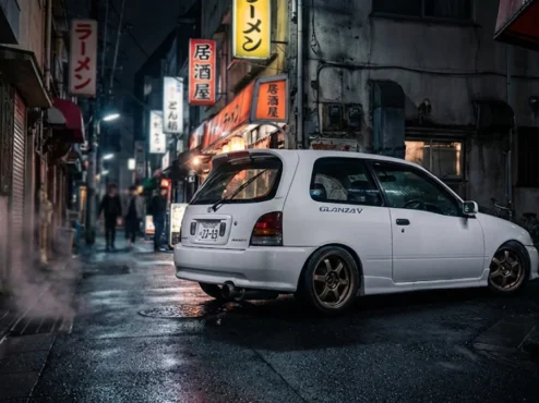 Toyota Starlet Glanza V branco rebaixado com rodas de liga leve em um cenário urbano japonês à noite.