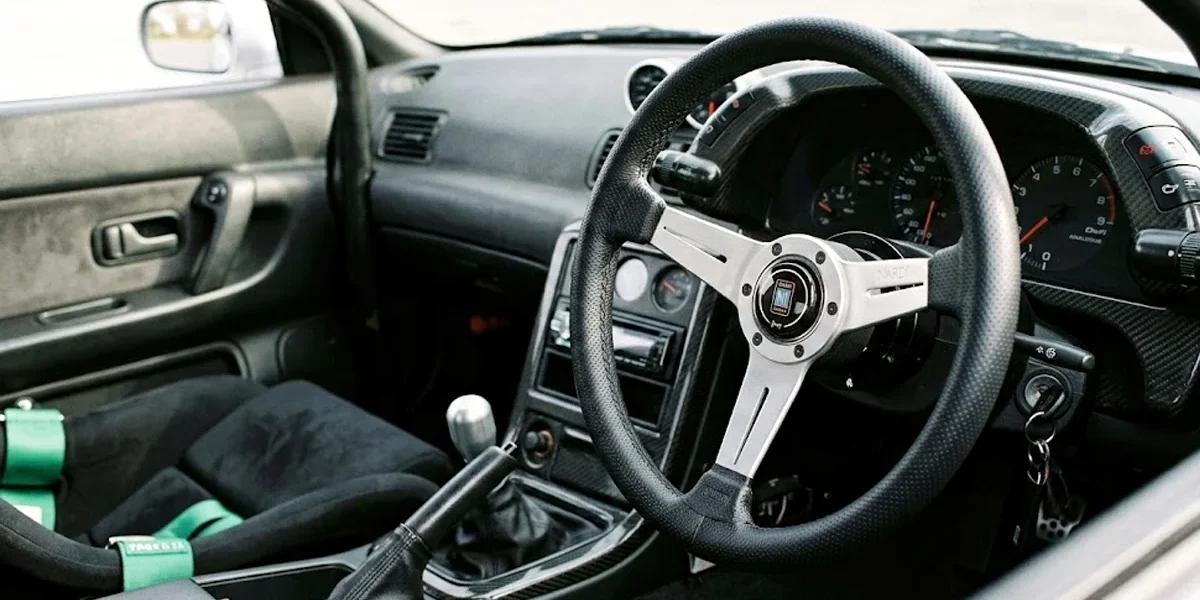 volante-esportivo-nardi-instalado-interior-carro-jdm Detalhe de um volante esportivo de marca Nardi instalado em um cockpit de carro customizado, mostrando o acabamento em couro.