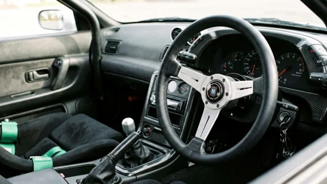 volante-esportivo-nardi-instalado-interior-carro-jdm Detalhe de um volante esportivo de marca Nardi instalado em um cockpit de carro customizado, mostrando o acabamento em couro.