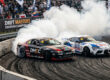 Dois carros de drift profissionais em uma batalha de proximidade extrema no Drift Masters, gerando muita fumaça em uma curva fechada.