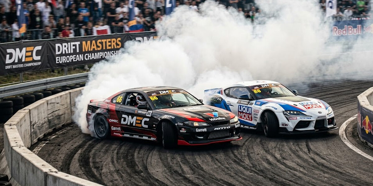 drift-masters-european-championship-tecnica-proximidade Dois carros de drift profissionais em uma batalha de proximidade extrema no Drift Masters, gerando muita fumaça em uma curva fechada.