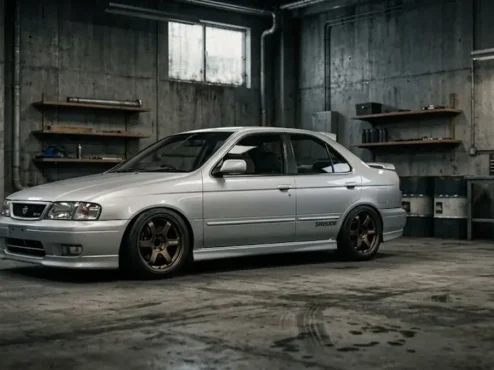 Nissan Sentra antigo prateado com modificação estilo JDM, rebaixado com rodas bronze em uma garagem industrial.