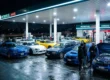 Encontro noturno de carros modificados estacionados sob as luzes brilhantes de um posto de gasolina em atmosfera gearhead.