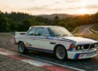 BMW 3.0 CSL Batmobile branco com faixas de corrida estacionado em curva de pista de circuito histórico ao pôr do sol.