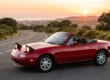 Mazda MX-5 Miata NA vermelho de primeira geração com os faróis escamoteáveis erguidos parado em uma estrada ao entardecer.