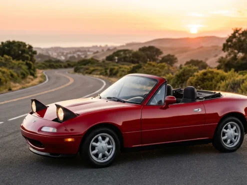 Mazda MX-5 Miata NA vermelho de primeira geração com os faróis escamoteáveis erguidos parado em uma estrada ao entardecer.