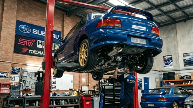 Subaru Impreza WRX azul clássico elevado em um elevador hidráulico dentro de uma oficina especializada em modificação automotiva.