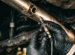Mecânico segurando sensor de oxigênio sonda lambda próximo ao escapamento de um carro em oficina.
