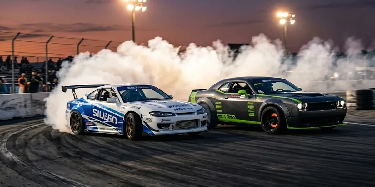 Dois carros de drift profissionais fazendo tandem em alta velocidade com pneus soltando fumaça densa na pista.