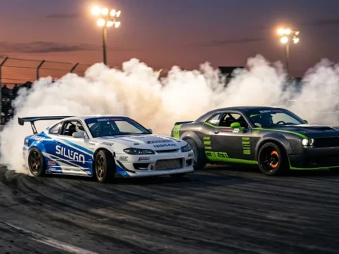 Dois carros de drift profissionais fazendo tandem em alta velocidade com pneus soltando fumaça densa na pista.