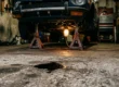 Poça de óleo escuro no chão de concreto de uma garagem com um carro esportivo desfocado ao fundo sobre cavaletes.