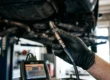 Mecânico automotivo inspecionando uma sonda lambda desgastada com um scanner de diagnóstico no fundo.