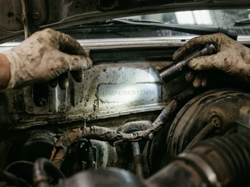 Mecânico iluminando com lanterna a numeração do chassi no cofre do motor durante vistoria cautelar automotiva.